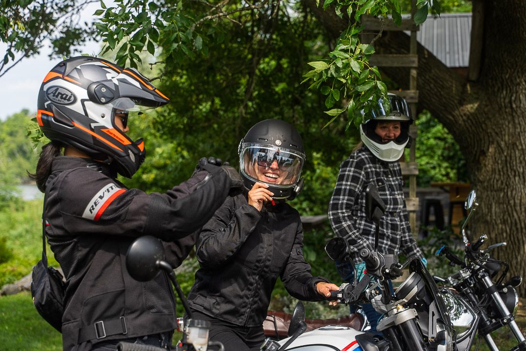 3 female motorcyle riders talking and laughing on their bikes under a shady green tree.
